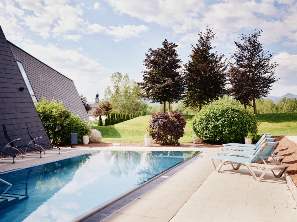 Außenbecken der AQUALUX Therme Fohnsdorf mit ruhiger Wasserfläche, Liegen am Beckenrand, gepflegter Gartenanlage und Bäumen vor sanfter Hügel- und Landschaftskulisse.