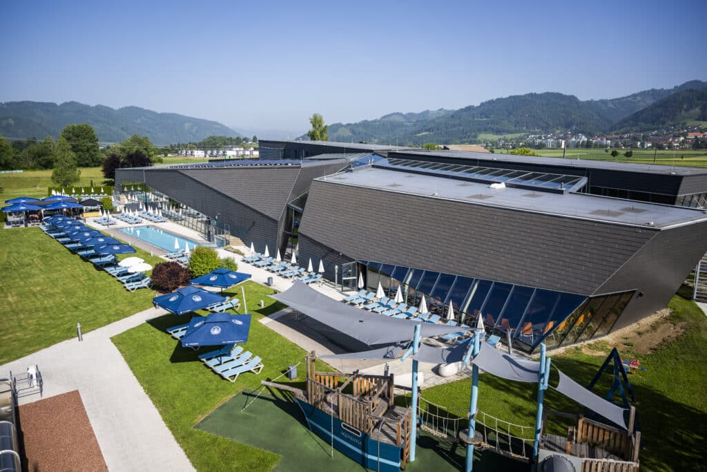 Luftaufnahme der AQUALUX Therme Fohnsdorf mit moderner Architektur, Außenbecken, Liegewiesen mit Sonnenschirmen, Spielbereich und umliegender grüner Landschaft vor Bergkulisse.